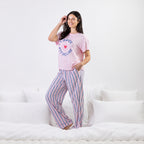 A woman modeling a pink pajama set consisting of a short-sleeve t-shirt with a blue and red graphic print and matching pink striped pajama pants, standing on a white bed.