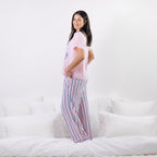 A woman stands on a white bed wearing a light pink pajama t-shirt and matching pajama pants with vertical blue and pink stripes.