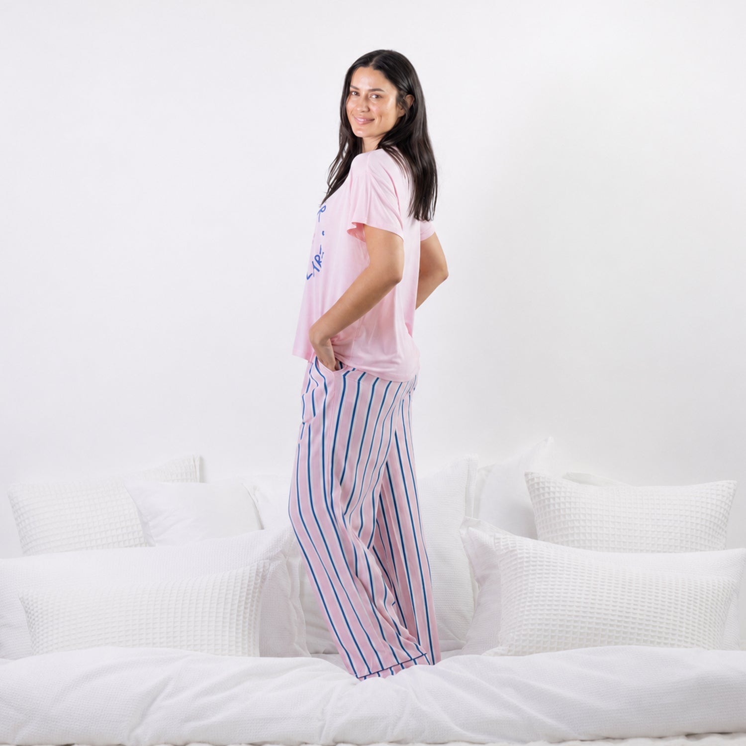 A woman stands on a white bed wearing a light pink pajama t-shirt and matching pajama pants with vertical blue and pink stripes.