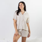 A woman modeling a matching short-sleeve button-up shirt and shorts pajama set in a cream and brown vertical stripe pattern, set against a clean white studio background.