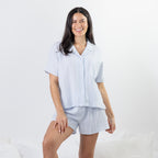 A woman modeling a light blue and white vertically striped short-sleeve button-up shirt and matching shorts pajama set against a clean white studio background.