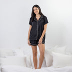 A woman stands on a bed wearing a black short-sleeve pajama set with white piping trim. The set includes a button-front top and matching shorts.