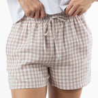 A close-up of a person wearing loose, gingham shorts in a natural and white check pattern with a matching drawstring waist. 