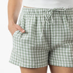 A close-up of a person wearing light green and white gingham check shorts with an elastic drawstring waistband and side pockets.