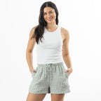 A woman modeling a white ribbed tank top and sage green and white gingham check shorts against a plain white background.