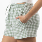 A close-up of a person wearing light green and white gingham check shorts with an elastic drawstring waist and side pockets, paired with a white ribbed top.