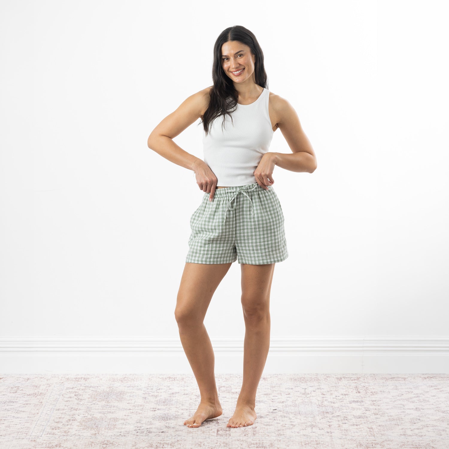A woman stands barefoot on a pale patterned rug against a white wall, wearing a white ribbed tank top and light green and white gingham drawstring shorts.