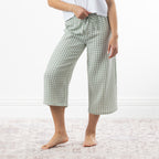 A person wearing sage green and white gingham capri pants with a drawstring waist, standing barefoot on a pale patterned rug against a white wall.