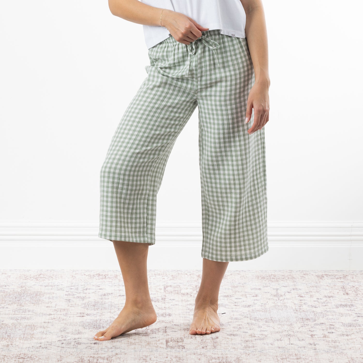 A person wearing sage green and white gingham capri pants with a drawstring waist, standing barefoot on a pale patterned rug against a white wall.