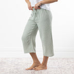 A person wearing sage green and white gingham capri pants with a drawstring waist, standing barefoot on a pale patterned rug against a white wall.