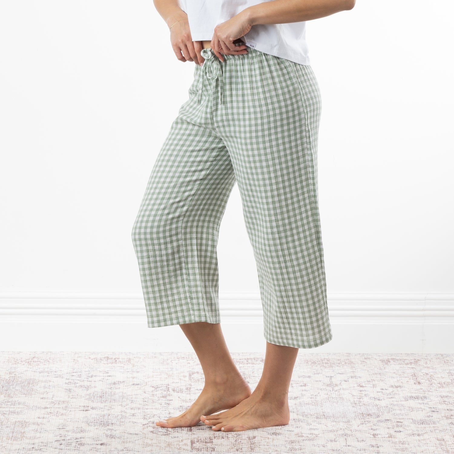 A person wearing sage green and white gingham capri pants with a drawstring waist, standing barefoot on a pale patterned rug against a white wall.