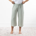 A person wearing sage green and white gingham capri pants with a drawstring waist, standing barefoot on a pale patterned rug against a white wall.