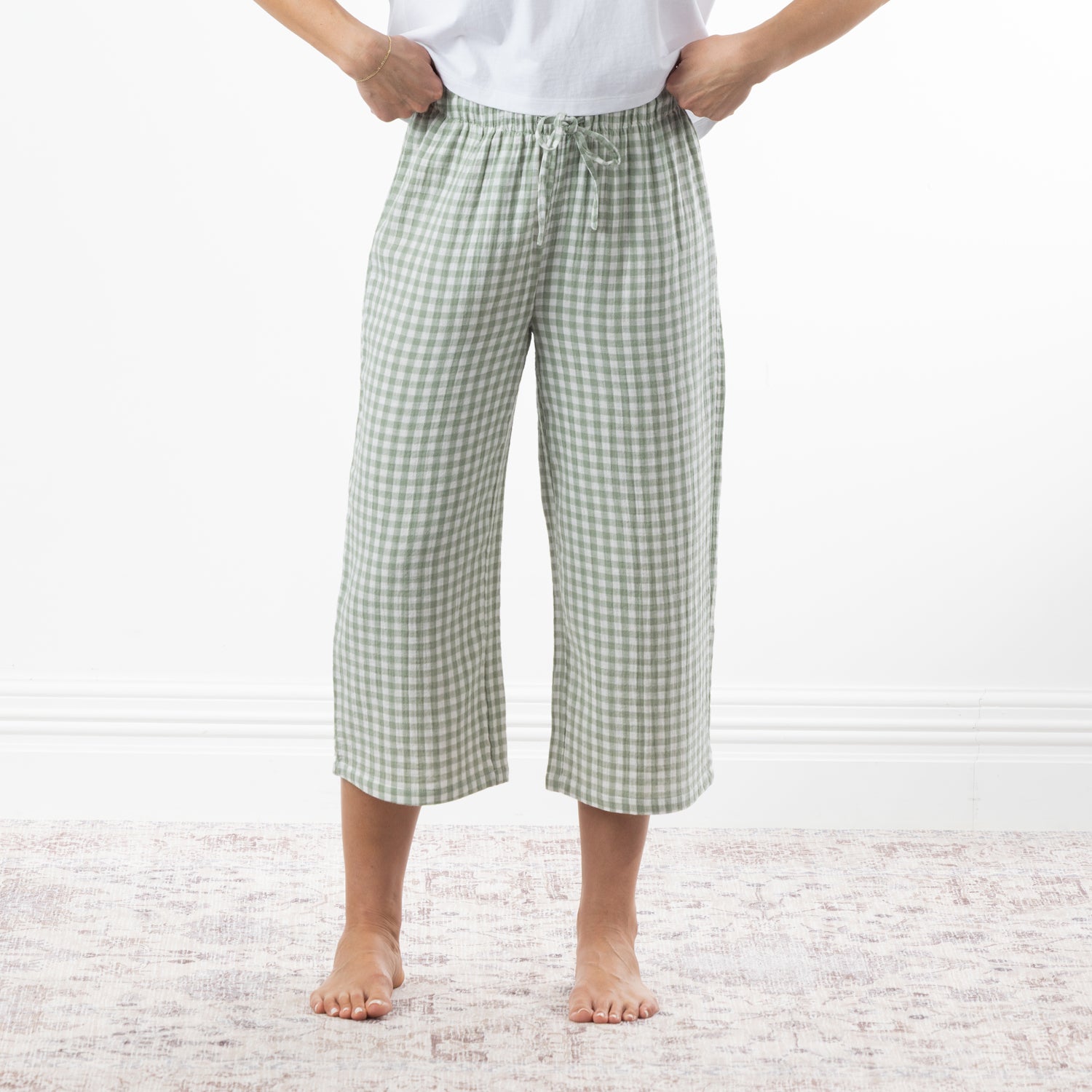 A person wearing sage green and white gingham capri pants with a drawstring waist, standing barefoot on a pale patterned rug against a white wall.
