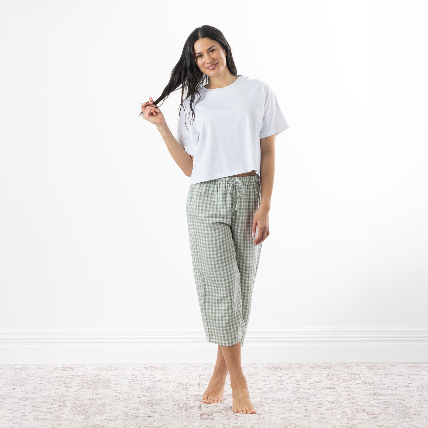 A woman stands barefoot in a bright room wearing a white short-sleeve crop top and sage-and-white gingham capri pants.