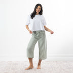 A woman stands barefoot in a bright room wearing a white short-sleeve crop top and sage-and-white gingham capri pants.