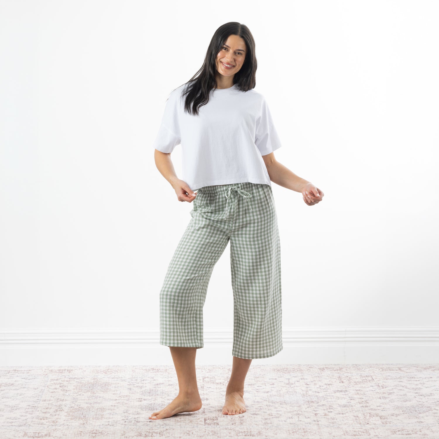 A woman stands barefoot in a bright room wearing a white short-sleeve crop top and sage-and-white gingham capri pants.