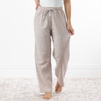 A person wearing loose, straight-leg pajama pants with a natural and white gingham check pattern, standing barefoot on a light patterned rug against a white wall.