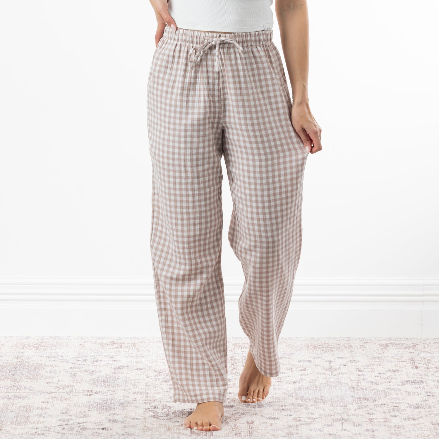 A person wearing loose, straight-leg pajama pants with a natural and white gingham check pattern, standing barefoot on a light patterned rug against a white wall.