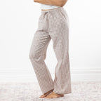 A person wearing loose, straight-leg pajama pants with a natural and white gingham check pattern, standing barefoot on a light patterned rug against a white wall.