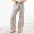 A person wearing loose, straight-leg pajama pants with a natural and white gingham check pattern, standing barefoot on a light patterned rug against a white wall.
