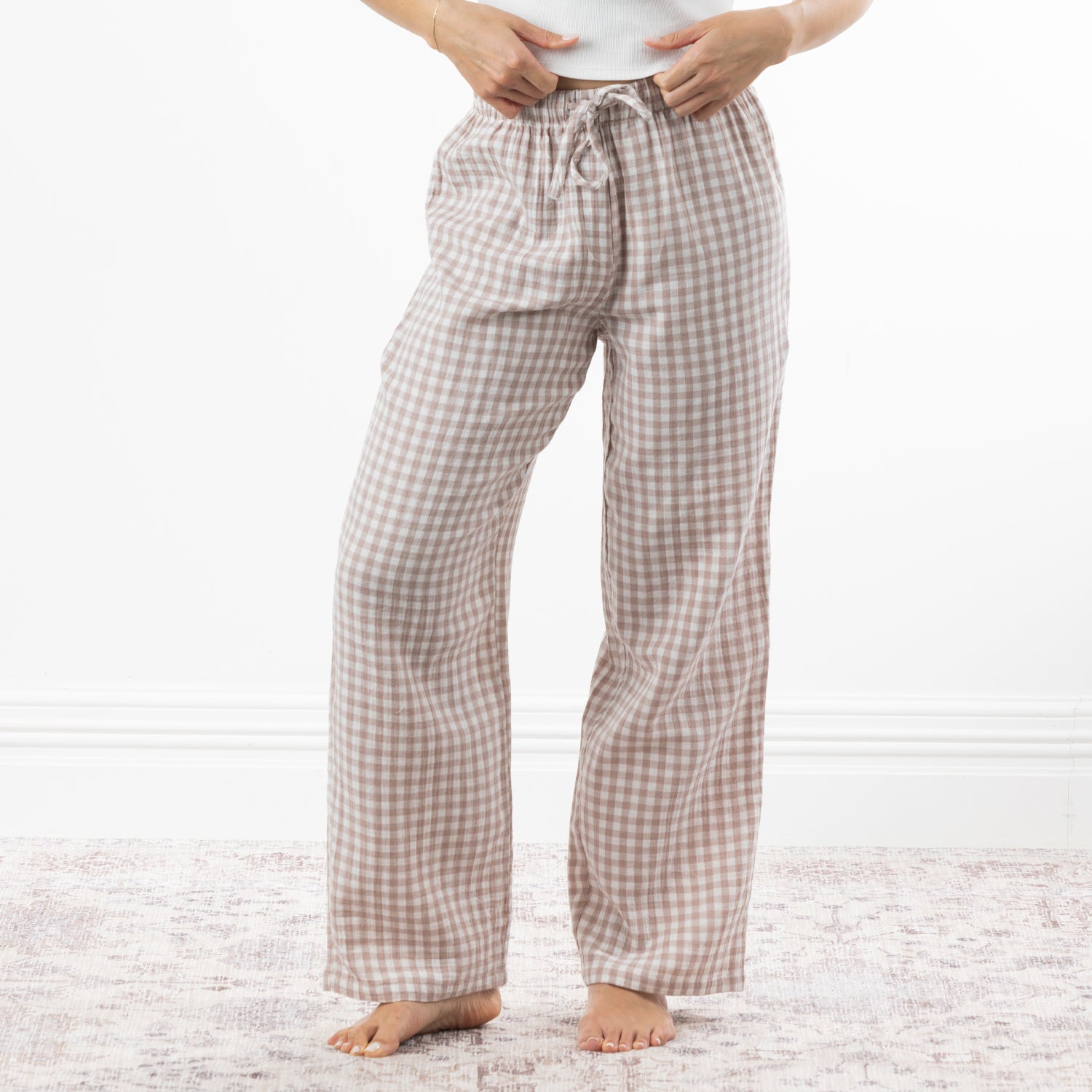 A person wearing loose, straight-leg pajama pants with a natural and white gingham check pattern, standing barefoot on a light patterned rug against a white wall.