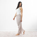 A woman stands barefoot in a bright white room wearing loose, gingham pajama pants in natural and white, paired with a white tank top.