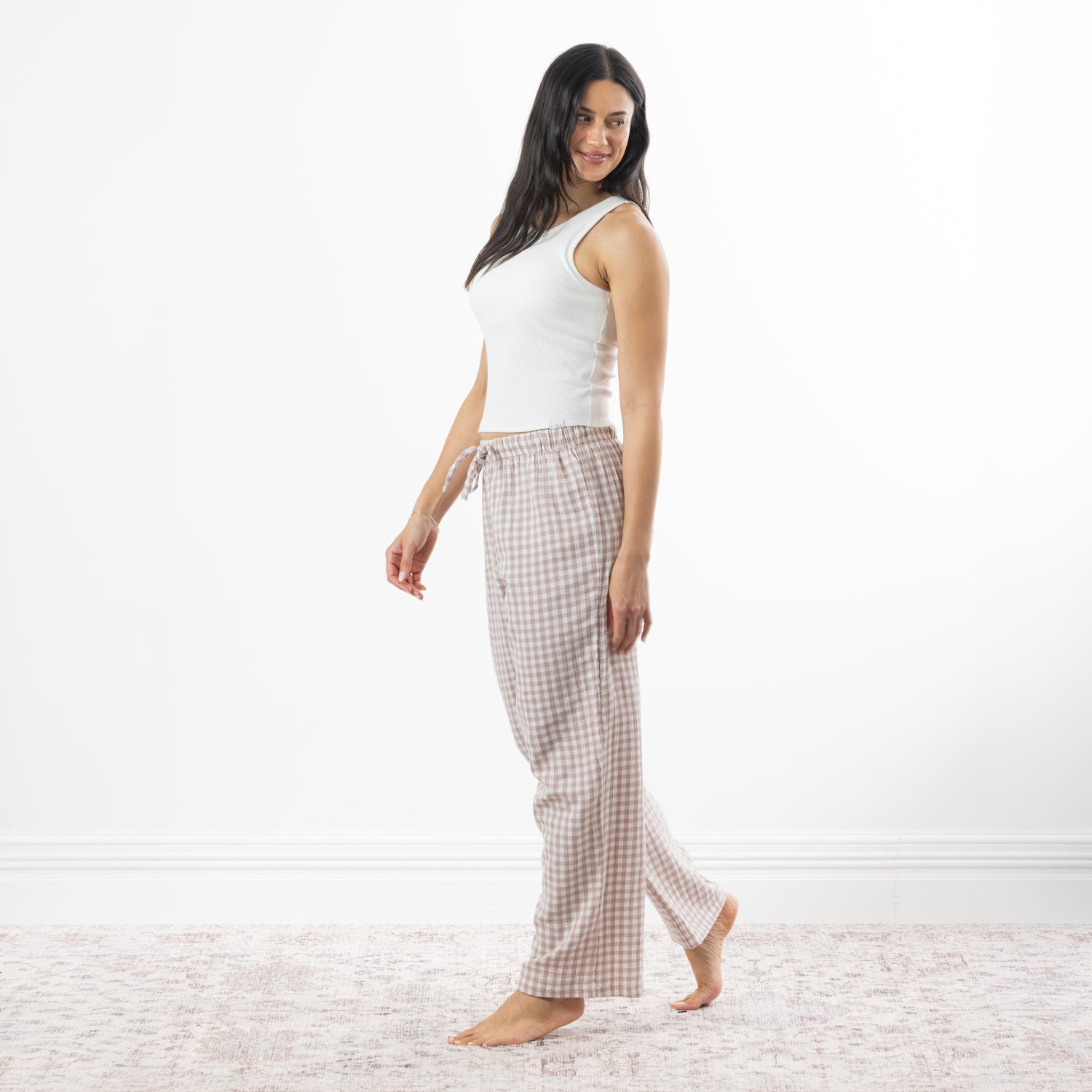 A woman stands barefoot in a bright white room wearing loose, gingham pajama pants in natural and white, paired with a white tank top.
