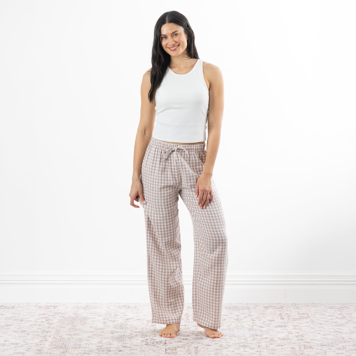 A woman stands barefoot in a bright white room wearing loose, gingham pajama pants in natural and white, paired with a white tank top.