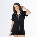 Woman wearing a black short-sleeve pyjama set with white contrast piping, button-front top and matching shorts, posed against a white studio background.