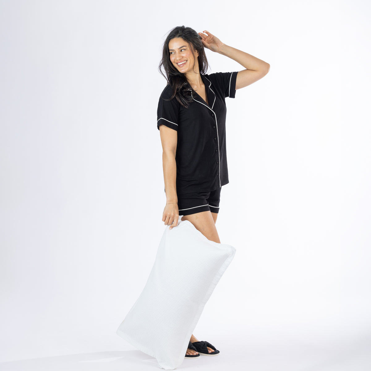 Woman wearing a black short pyjama set with white piping, posed in a relaxed side stance while holding a white pillow.