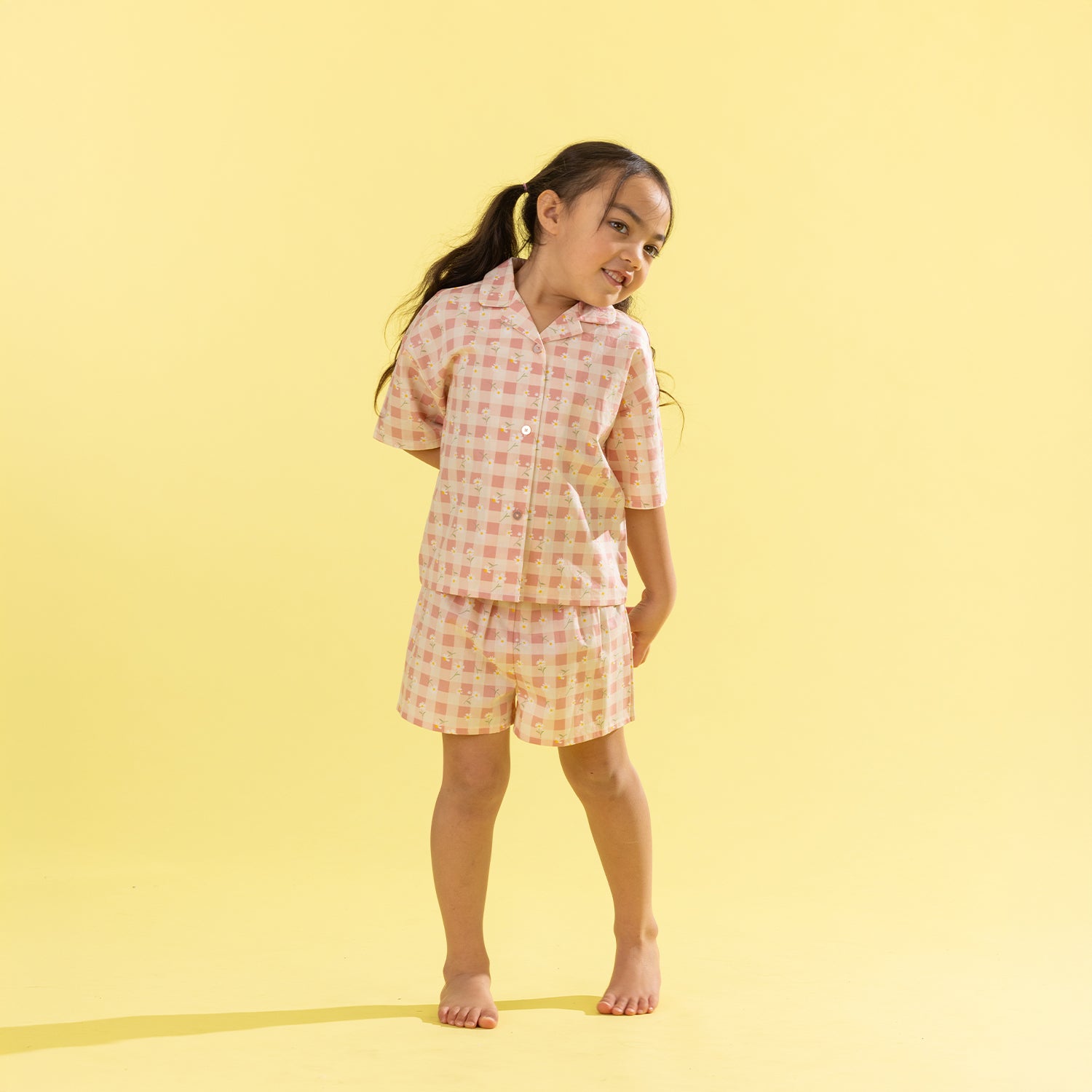 A young girl with her hair in a ponytail stands barefoot against a solid pale yellow background, wearing a two-piece pajama set with a pink and white gingham pattern.