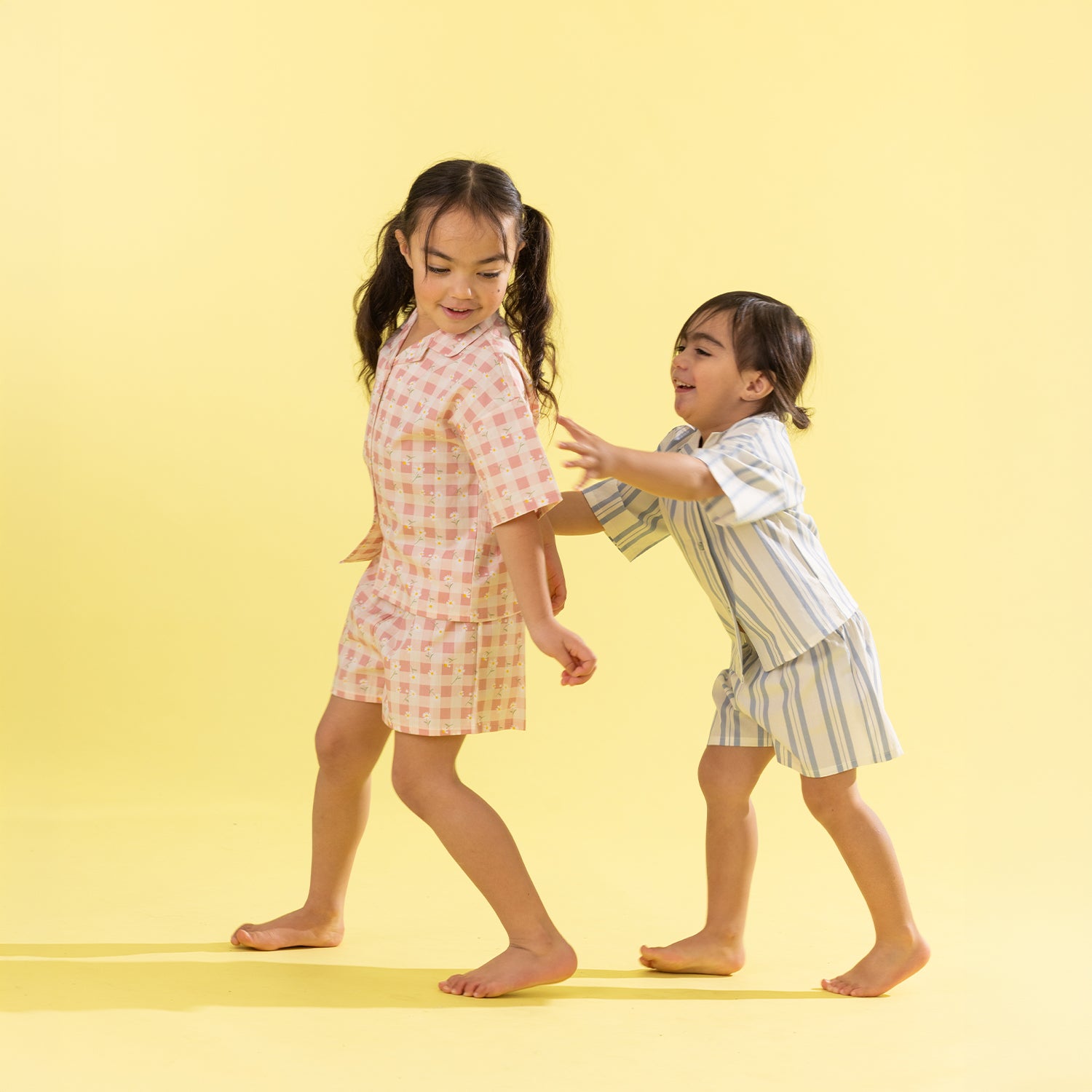 Two young girls playfully dancing in matching short pajama sets against a solid pale yellow background. The girl on the left wears a pink and white gingham check set, and the girl on the right wears a white set with thin blue vertical stripes.