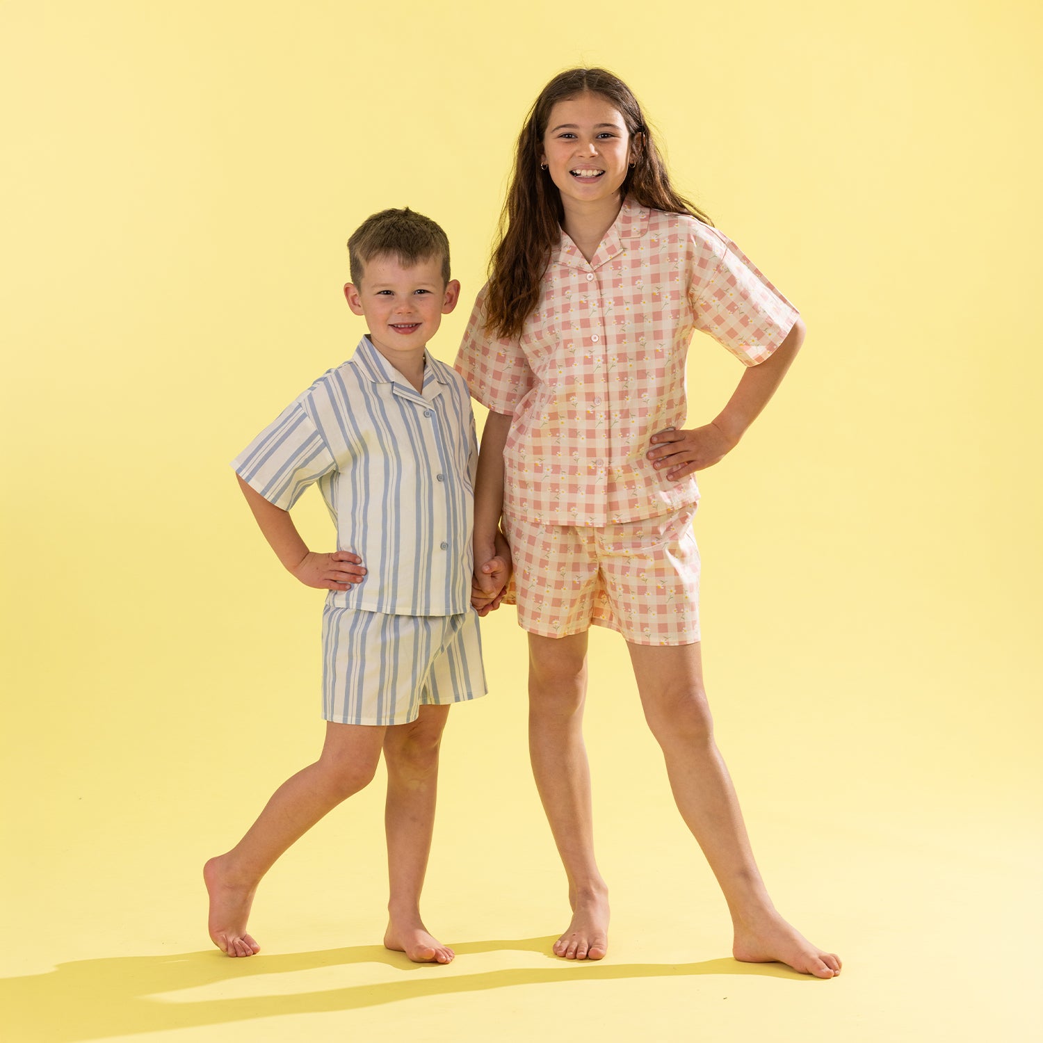 Two children modeling matching short-sleeve pajama sets against a solid pale yellow background. The boy wears a light blue and white vertically striped pajama shirt and shorts set, while the girl wears a pale pink and white gingham pajama shirt and shorts set.
