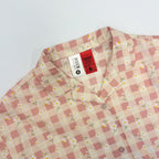 A close-up of a children's collared button-up shirt with a pink and cream gingham check pattern and a small white daisy print. The brand tag 'KIDS' is visible inside the collar.