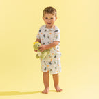 A young boy stands barefoot against a solid pale yellow background, wearing a matching two-piece pajama set with a white base and a colorful dinosaur print. He is holding a green plush toy.