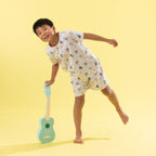 A young boy wearing a white short-sleeve pajama set with a colorful dinosaur print is balancing on one leg while holding a mint green ukulele against a solid yellow studio background.