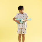 A young boy stands against a solid pale yellow background, smiling and playing a light blue ukulele. He is wearing a matching two-piece pajama set with a white base and a colorful cartoon print of characters and flowers.
