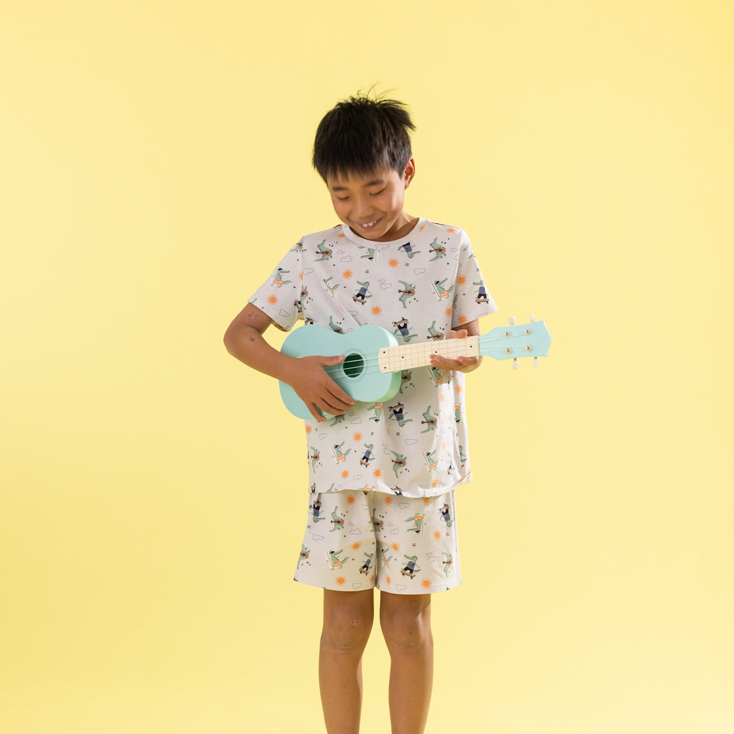A young boy stands against a solid pale yellow background, smiling and playing a light blue ukulele. He is wearing a matching two-piece pajama set with a white base and a colorful cartoon print of characters and flowers.