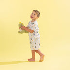 A young boy wearing a white two-piece pajama set with a colorful cartoon dinosaur pattern, standing barefoot on a solid pale yellow background while holding a green plush dinosaur toy.