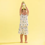 A smiling young child stands barefoot against a solid pale yellow background, holding a plush green toy above their head. The child is wearing a matching two-piece pajama set with a white base and a playful print of green leaves, black silhouettes, and small orange dots.