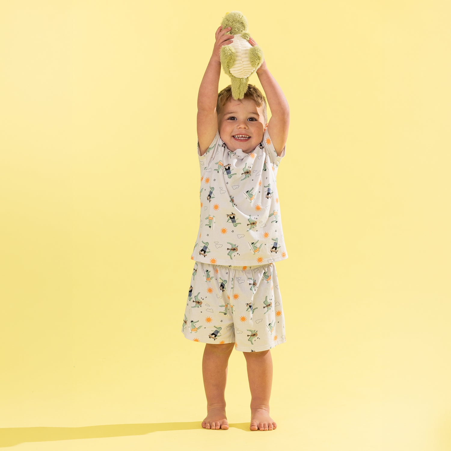 A smiling young child stands barefoot against a solid pale yellow background, holding a plush green toy above their head. The child is wearing a matching two-piece pajama set with a white base and a playful print of green leaves, black silhouettes, and small orange dots.
