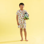 A young boy stands barefoot against a solid pale yellow background, wearing a matching short-sleeve pajama top and shorts set with a colorful bird print. He is holding a folded light green towel.