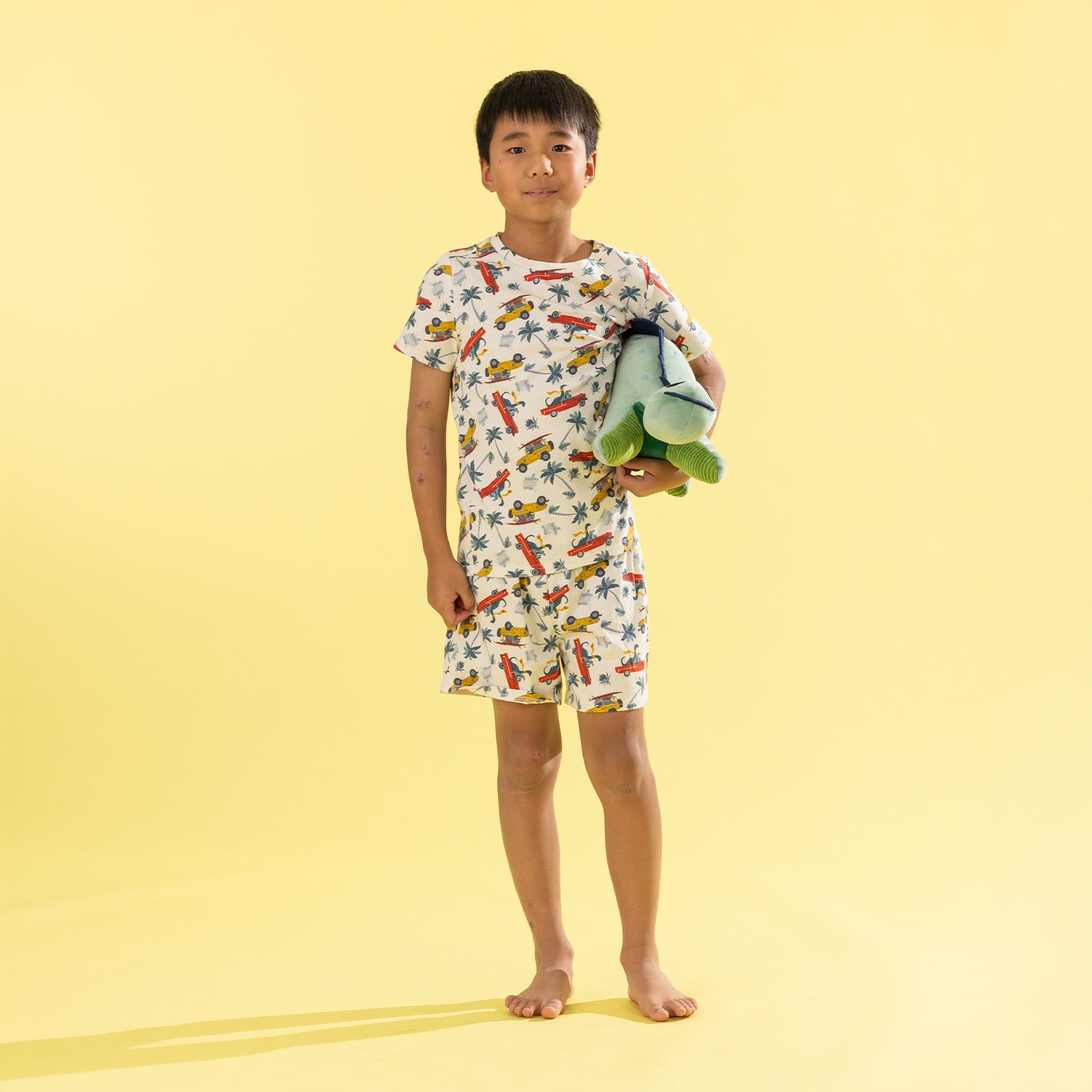 A young boy stands barefoot against a solid pale yellow background, wearing a matching short-sleeve pajama top and shorts set with a colorful bird print. He is holding a folded light green towel.