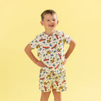 A young boy modeling a matching two-piece pajama set with a tropical car and palm tree print, posed against a solid pale yellow background.