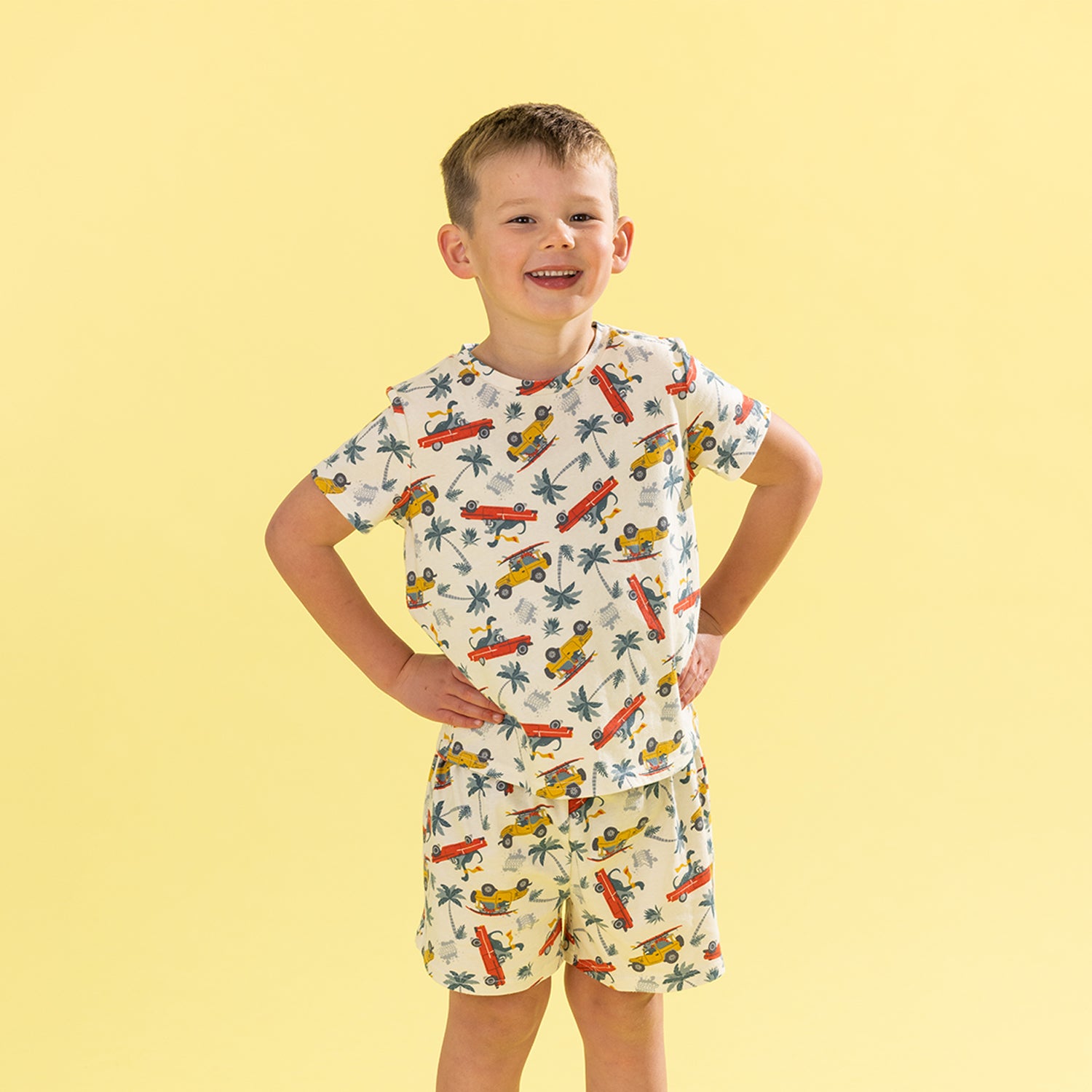 A young boy modeling a matching two-piece pajama set with a tropical car and palm tree print, posed against a solid pale yellow background.