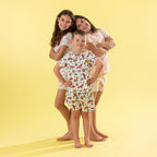 Three children modeling matching short-sleeve pajama sets against a solid pale yellow studio background. The set features a white base with a colorful pattern of toy cars, airplanes, and stars.