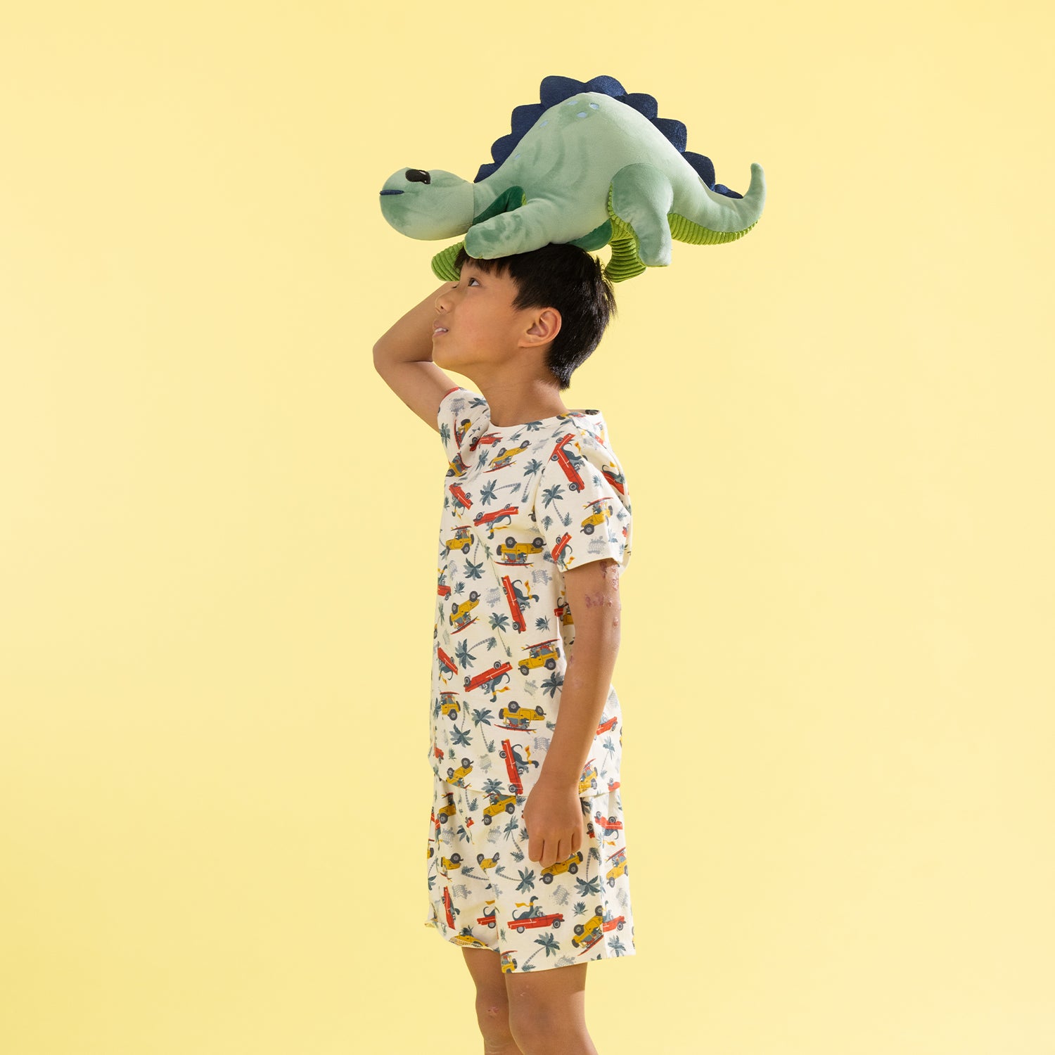 A child wearing a white short-sleeve dress with a colorful dinosaur and vehicle print, balancing a green plush dinosaur on their head against a solid pale yellow background.