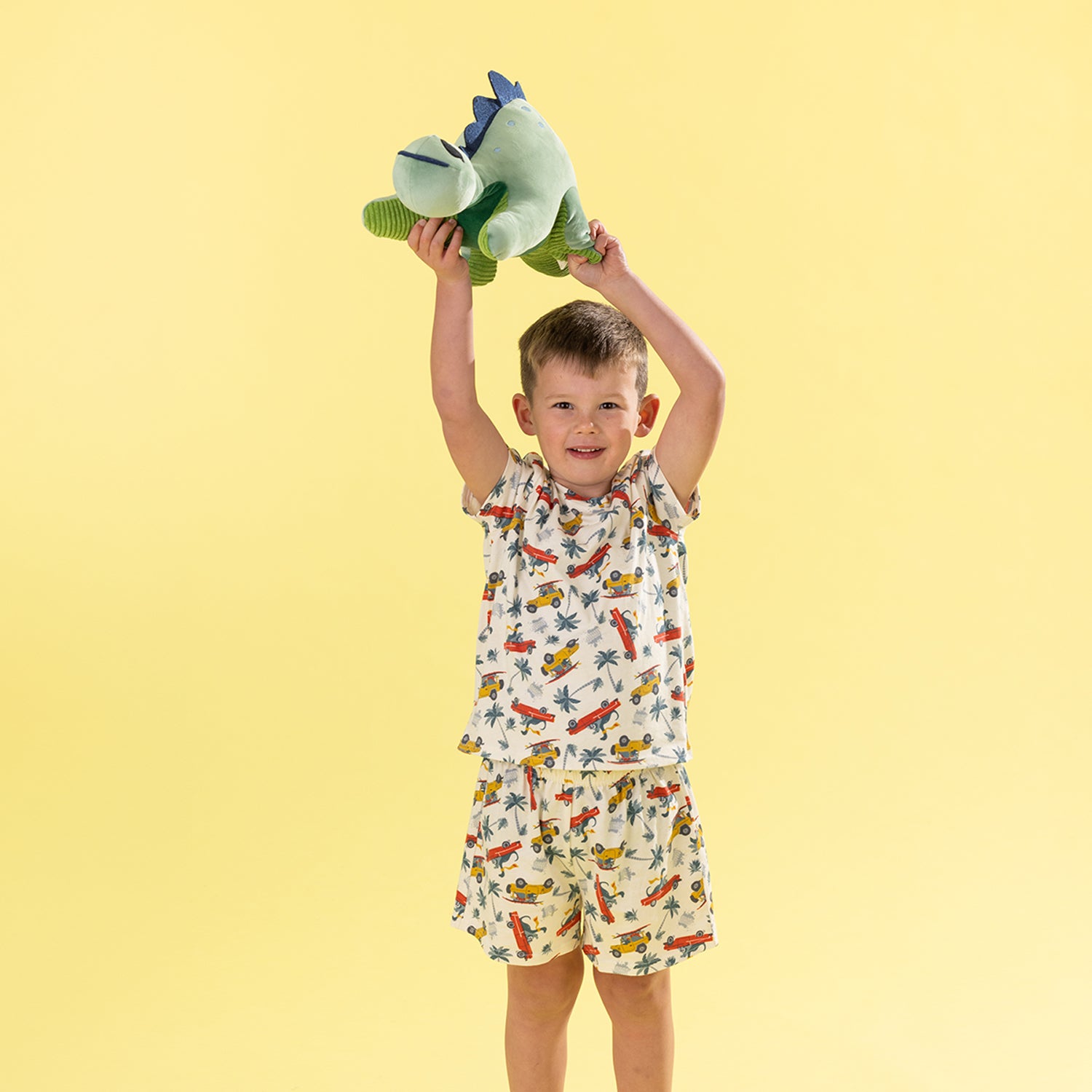 A young boy stands against a solid pale yellow background, holding a green plush dinosaur toy above his head. He is wearing a matching two-piece pajama set with a white base and a colorful print of airplanes, rockets, and stars.