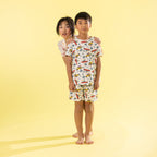 Two children modeling matching short-sleeve pajama sets against a solid pale yellow studio background. The boy in front wears a white pajama set with a colorful print of cars, airplanes, and palm trees, while the girl behind him wears a matching set with a different tropical print.