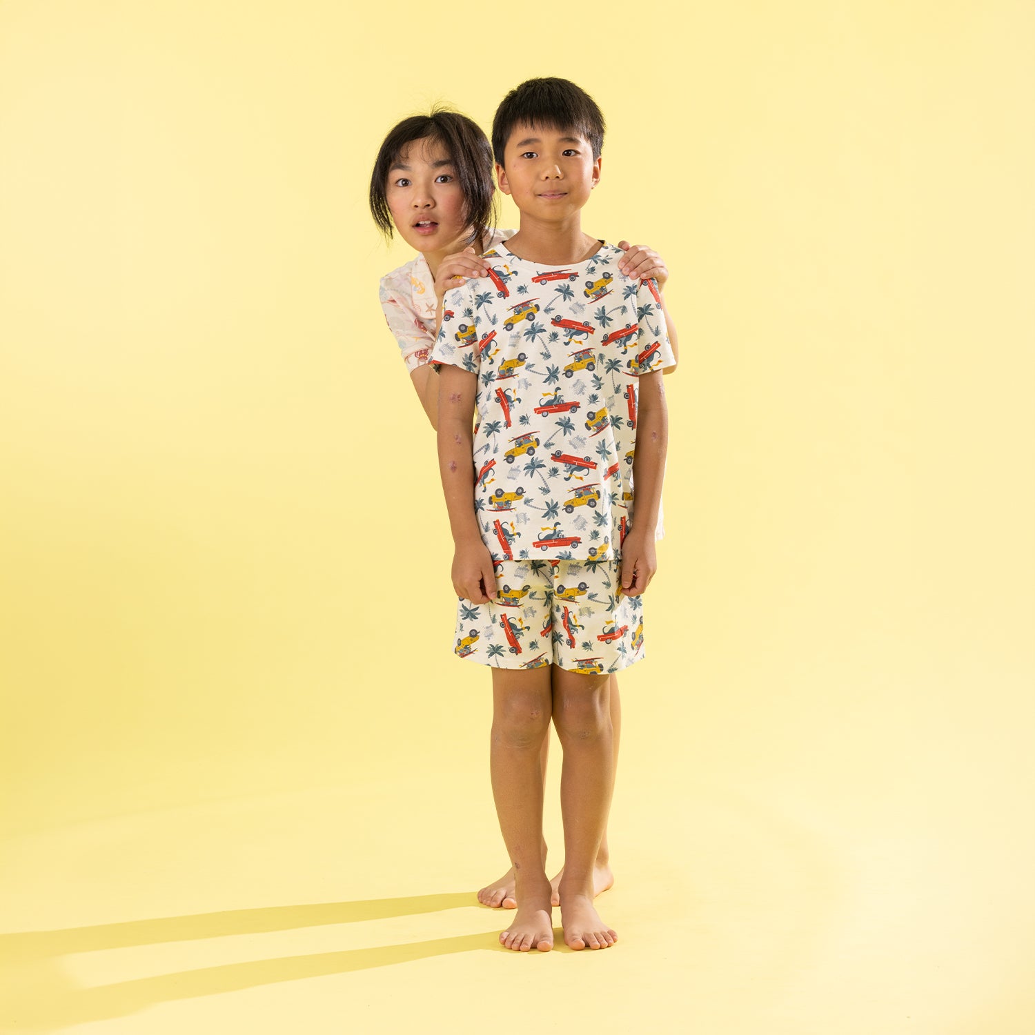 Two children modeling matching short-sleeve pajama sets against a solid pale yellow studio background. The boy in front wears a white pajama set with a colorful print of cars, airplanes, and palm trees, while the girl behind him wears a matching set with a different tropical print.